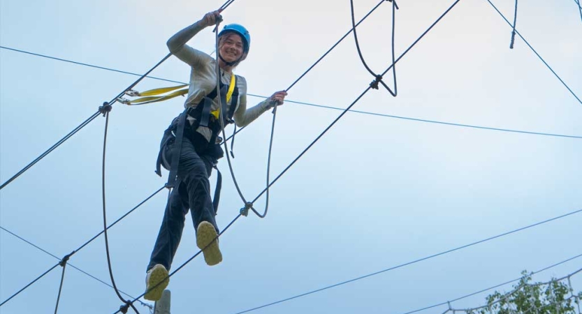 A person wearing safety gear and secured by ropes balances on an obstacle on a high ropes course. 